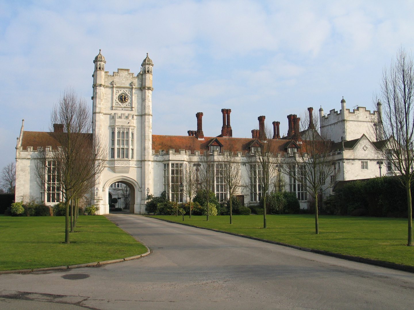 A grand white stately home with a tower, rows of regular windows and red tiled chimneys. A drive flanked by young trees leads up to the house.