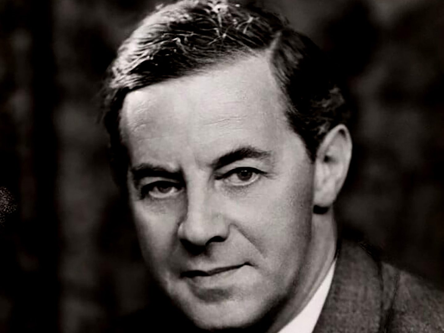 A historic black and white photo of a man looking at the camera. He has a side parting, a serious expression and is wearing a jacket.