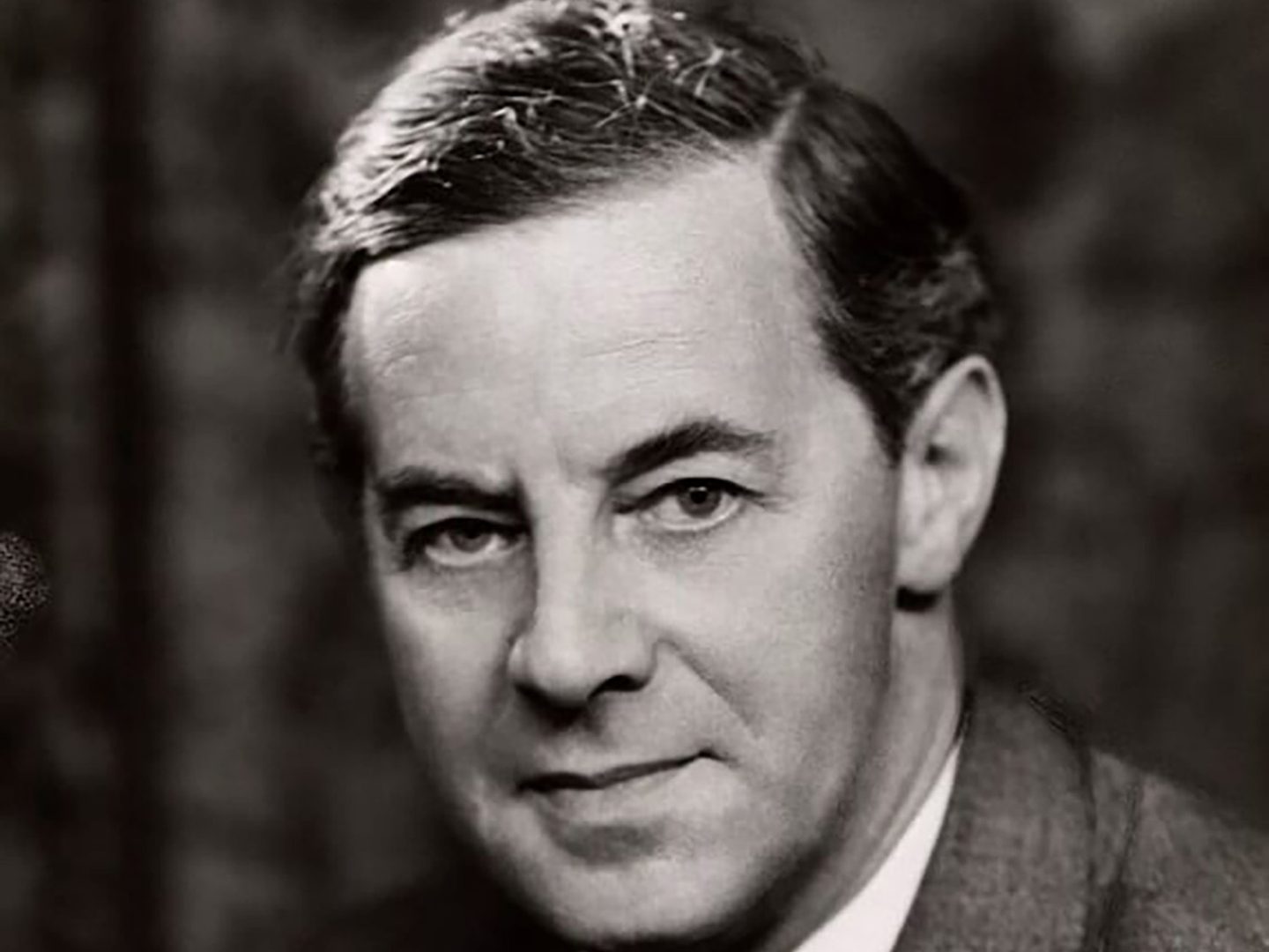 A historic black and white photo of a man looking at the camera. He has a side parting, a serious expression and is wearing a jacket.