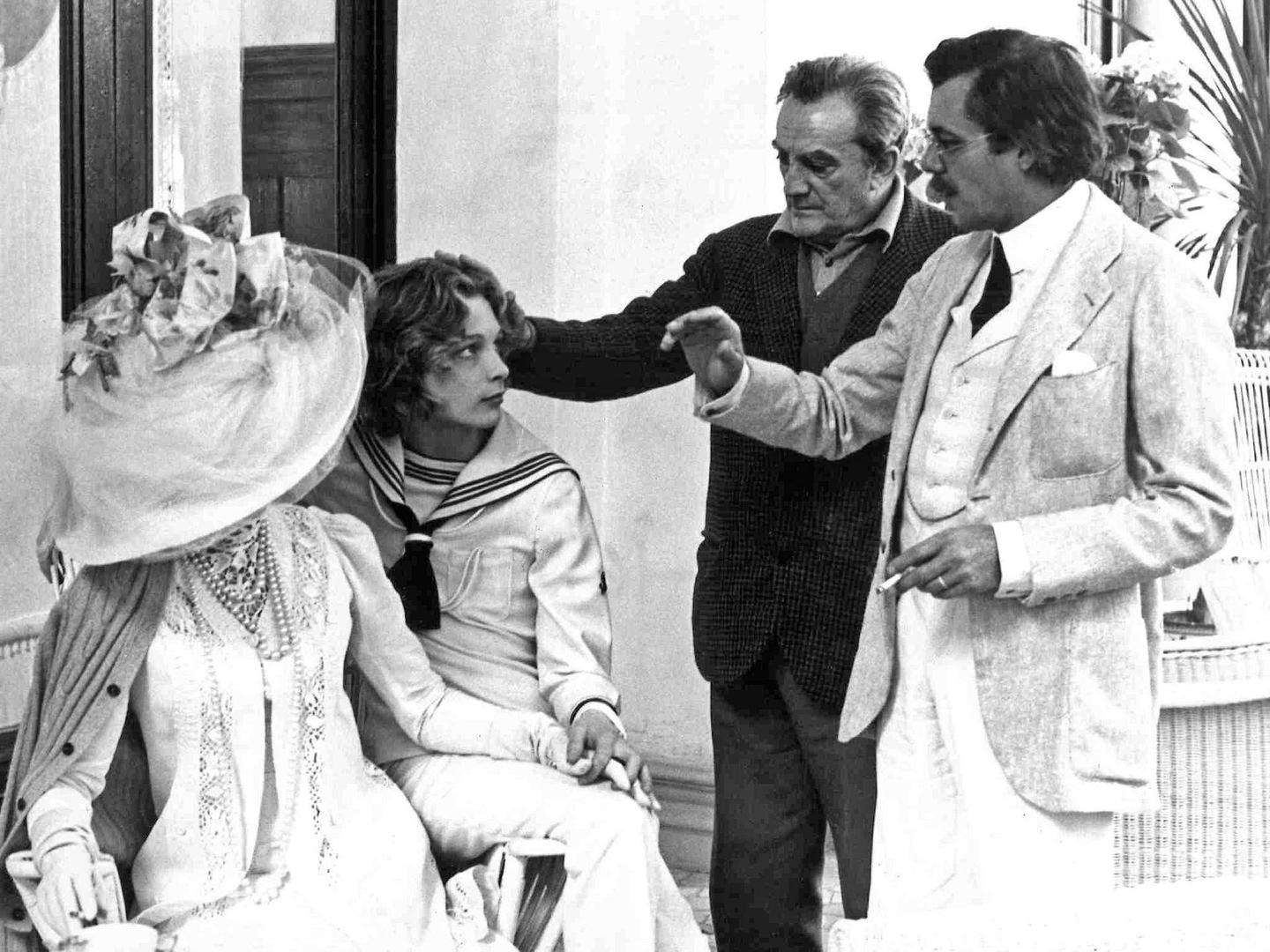 A historic photo of a group of 4 people. Dirk Bogarde stands to the right in a light-coloured suit. Behind him is a second man, while a woman in a large hat and a young man sit to the left of the image.