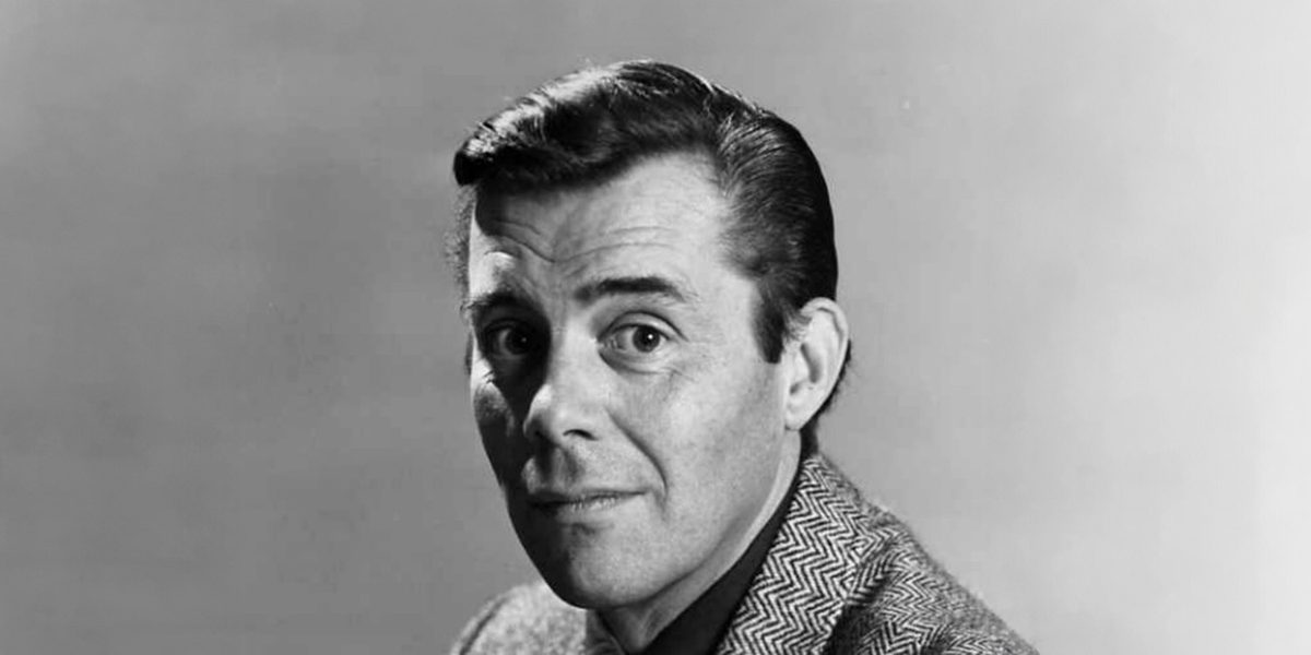 A head-shot of Dirk Bogarde staring straight at the camera. His dark hair is slicked back, he has raised his left eyebrow and he is wearing a tweed jacket.