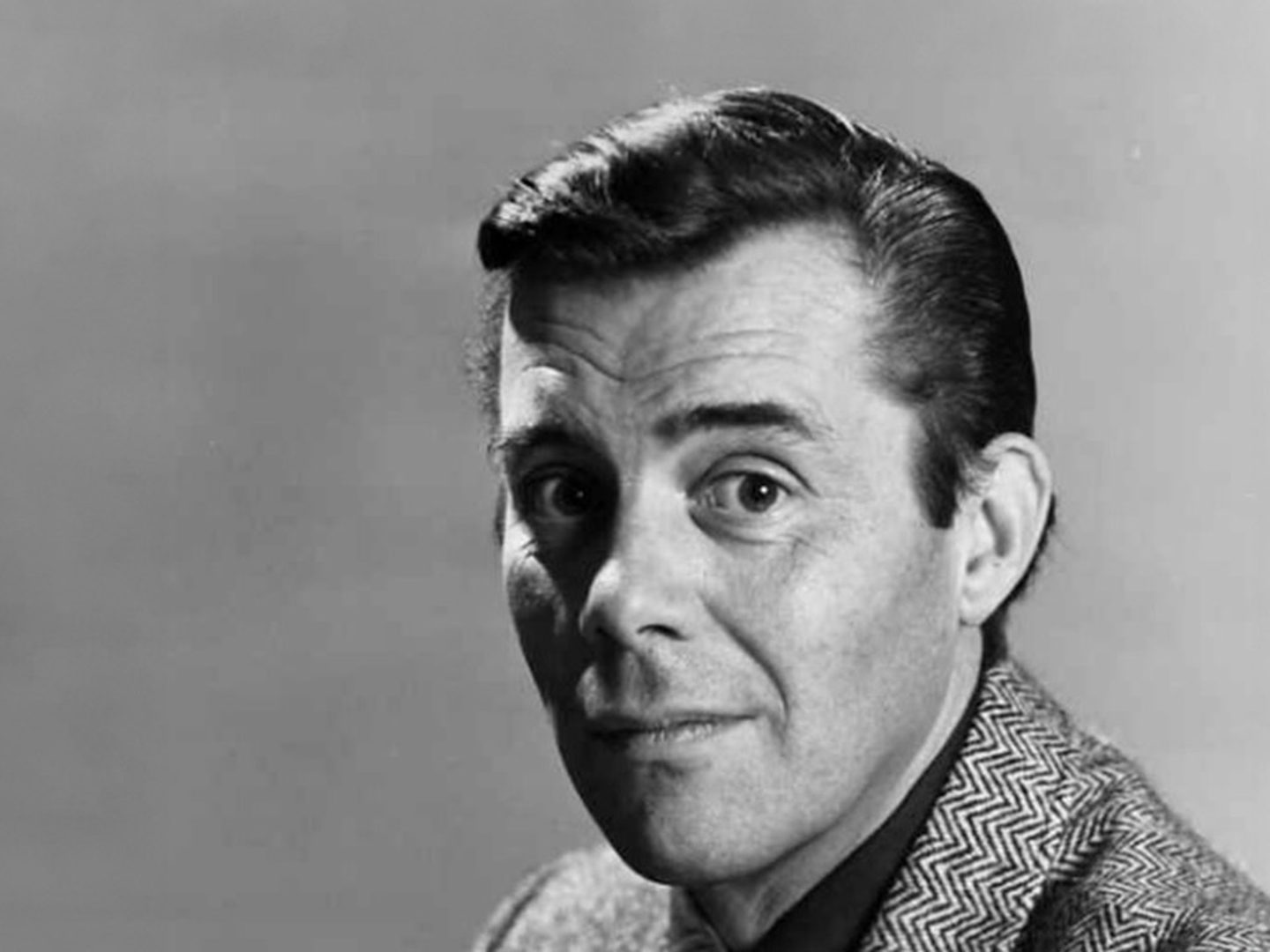 A head-shot of Dirk Bogarde staring straight at the camera. His dark hair is slicked back, he has raised his left eyebrow and he is wearing a tweed jacket.