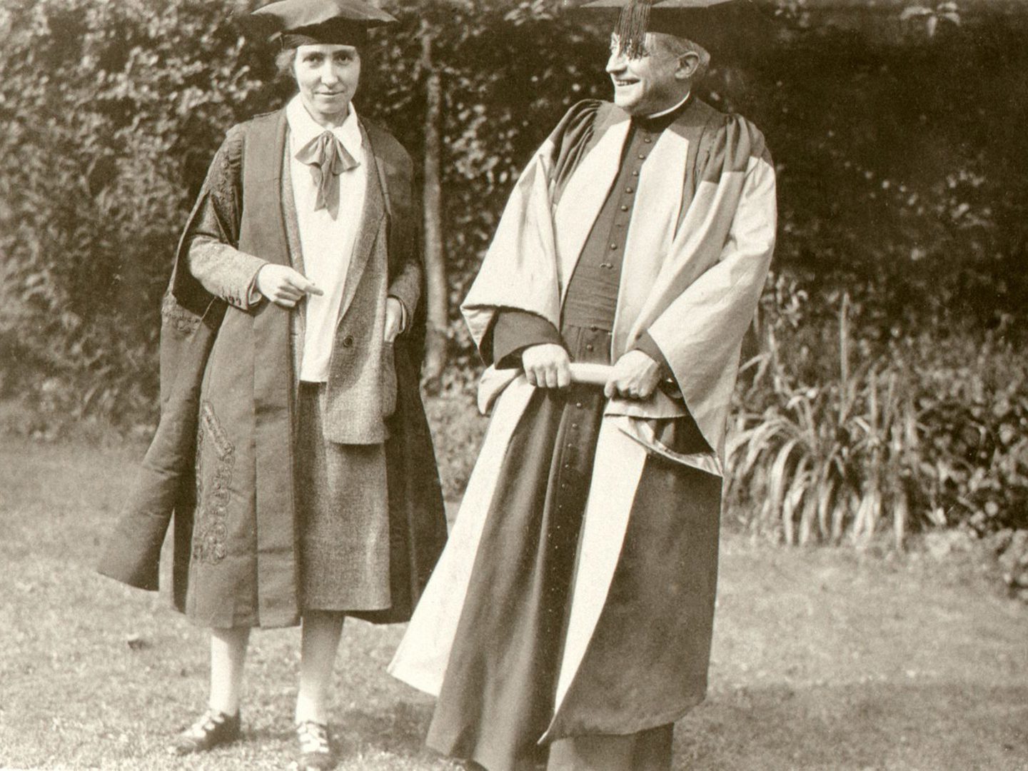A black and white historic photo of a man and a woman wearing academic caps and gowns, standing next to each other in a garden.