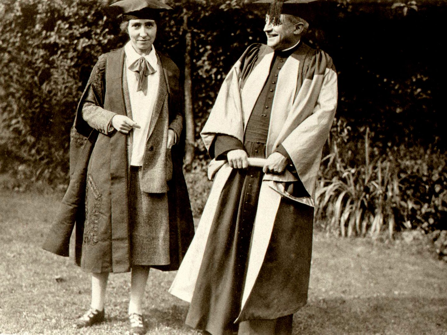 A black and white historic photo of a man and a woman wearing academic caps and gowns, standing next to each other in a garden.