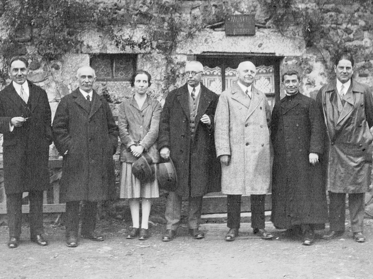 A black and white historic photo of a row of 6 men and 1 woman standing in front of a foliage-covered building.