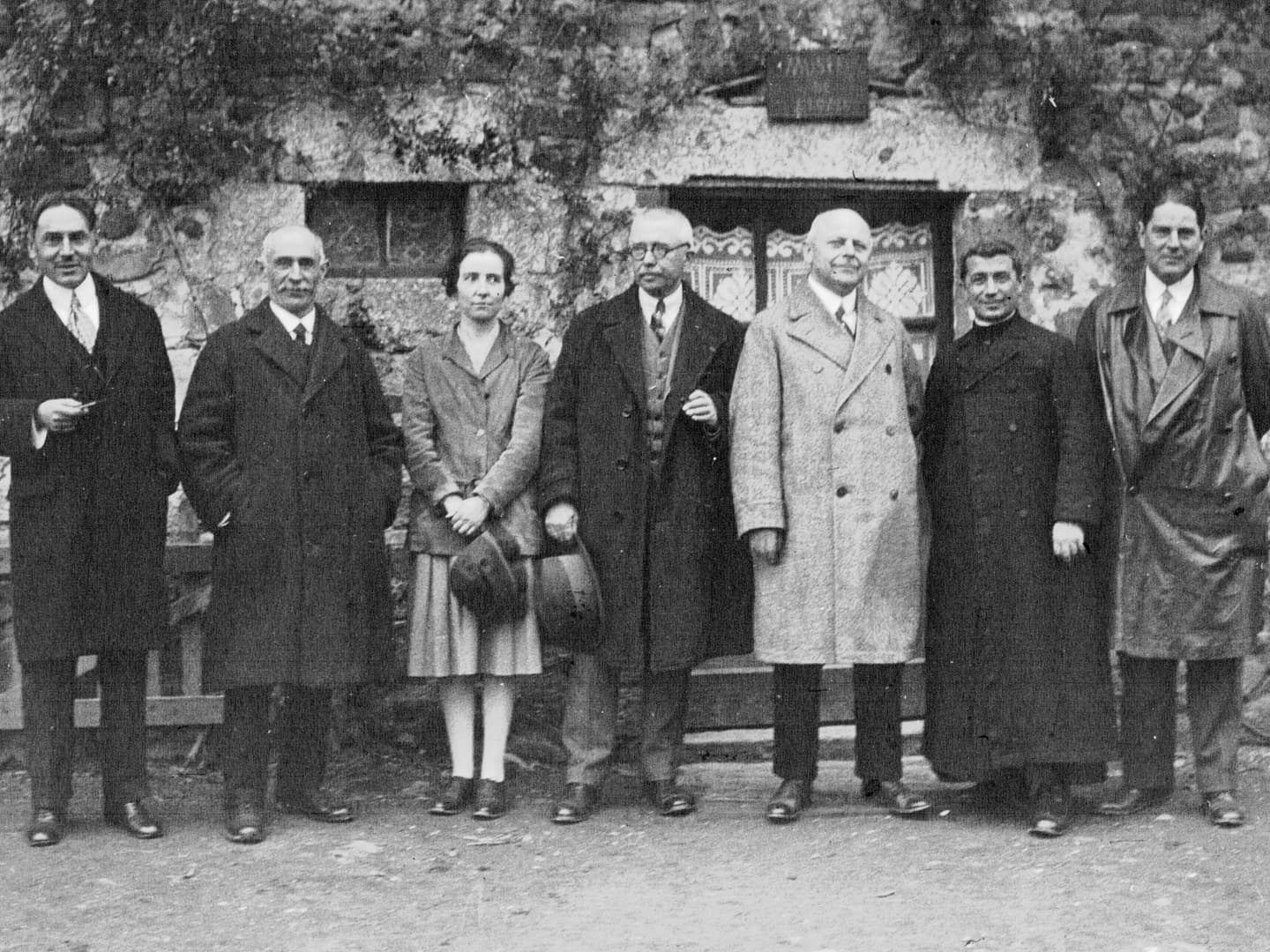 A black and white historic photo of a row of 6 men and 1 woman standing in front of a foliage-covered building.