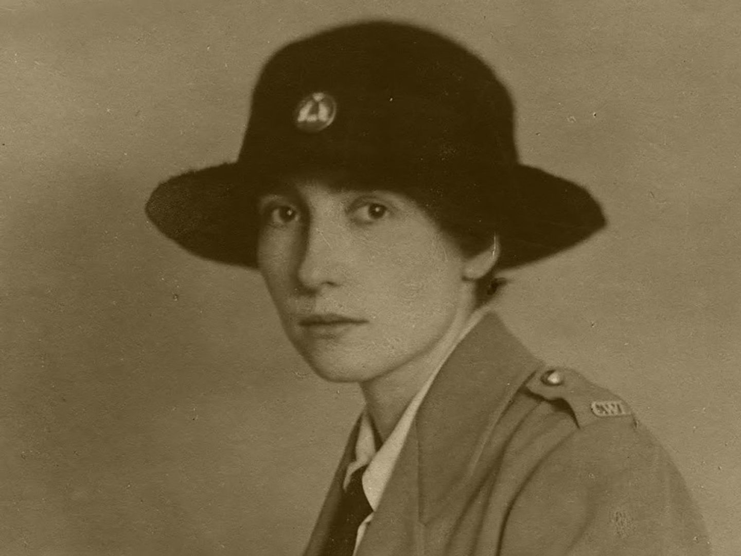 A sepia historic photo of a young woman in uniform, wearing a wide-brimmed black hat and looking at the viewer.