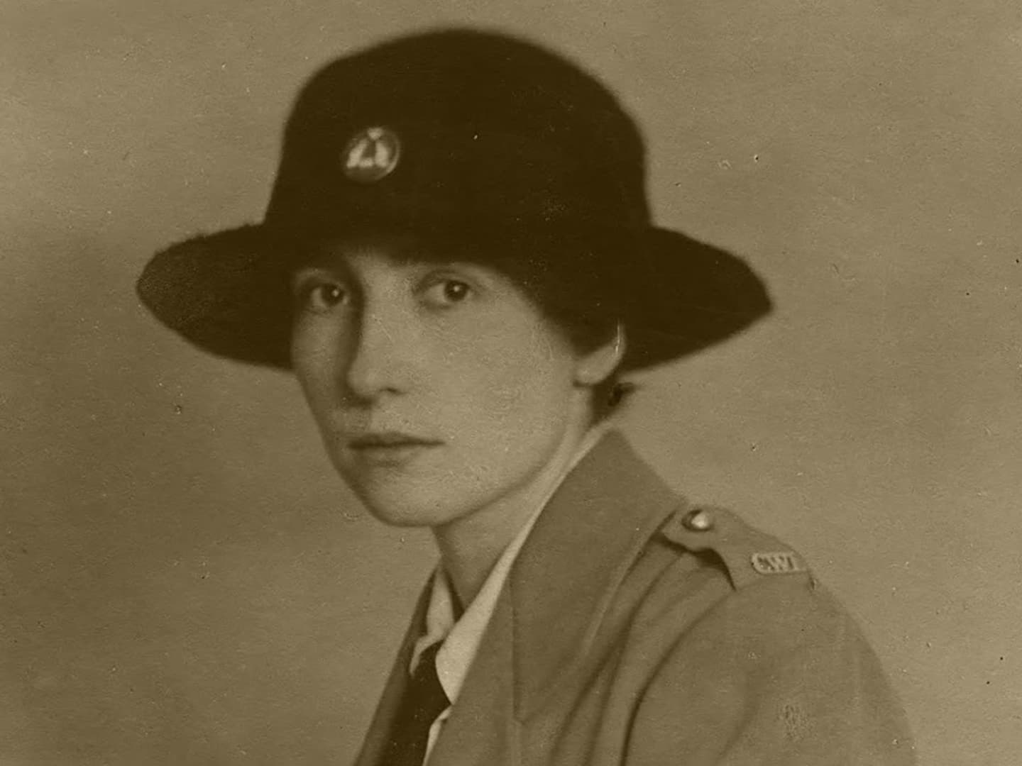 A sepia historic photo of a young woman in uniform, wearing a wide-brimmed black hat and looking at the viewer.