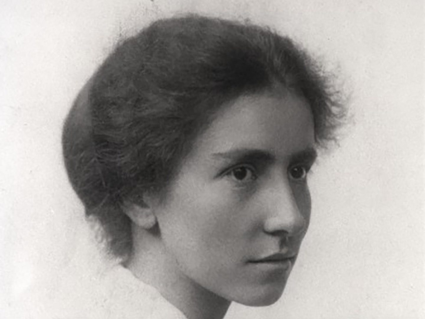 A black and white historic photo of a young woman looking past the viewer. Her frizzy hair is pulled back from her face and she is wearing a white top.
