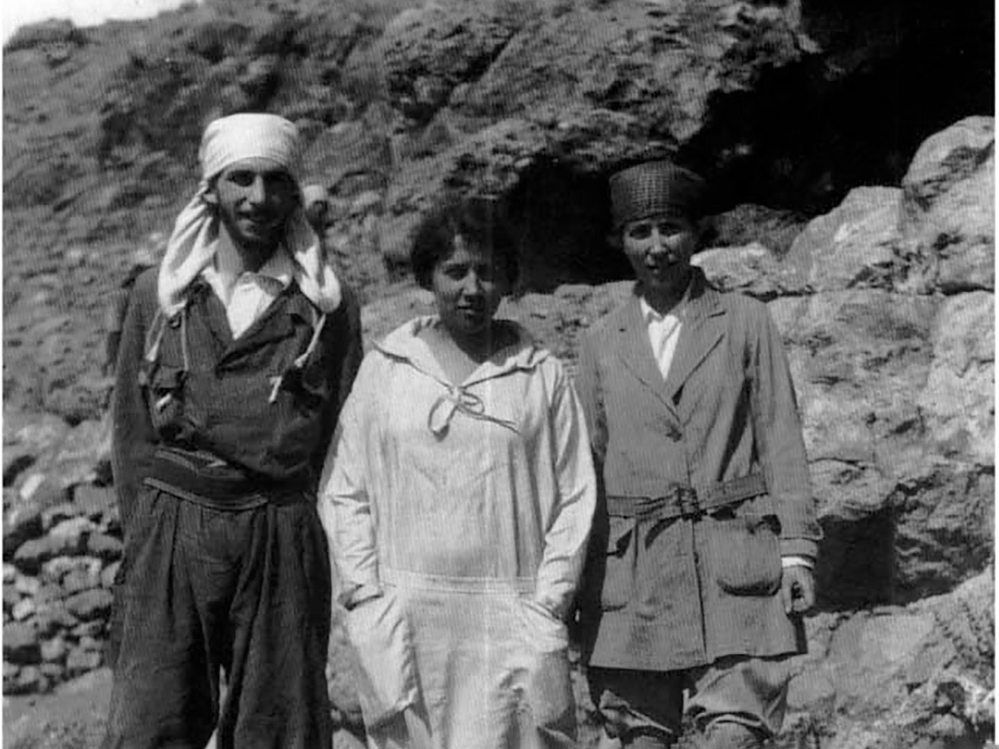A black and white historic photo of 2 women and a man wearing practical clothing and standing in a rocky landscape.