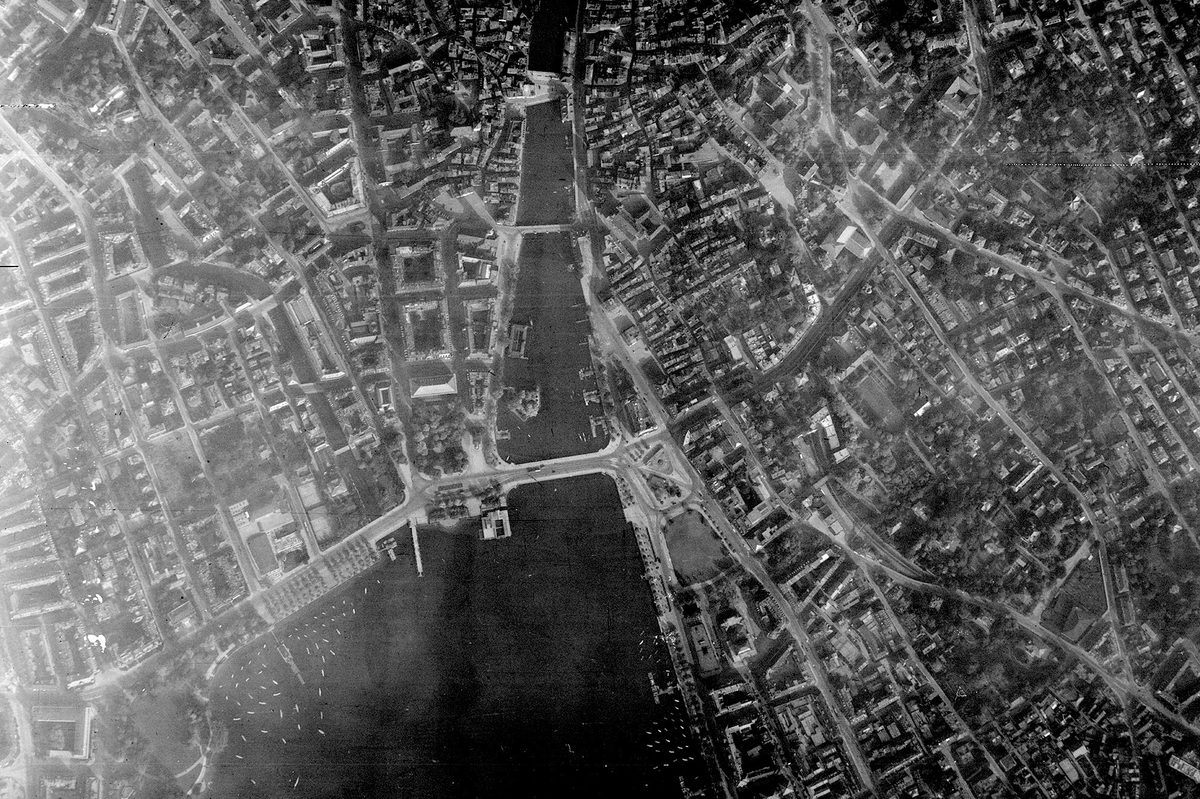A historic black and white vertical aerial photo of a large city straddling a river. 3 bridges cross the river, which runs into a lake at the bottom edge of the image. The city's streets and buildings are clearly visible.
