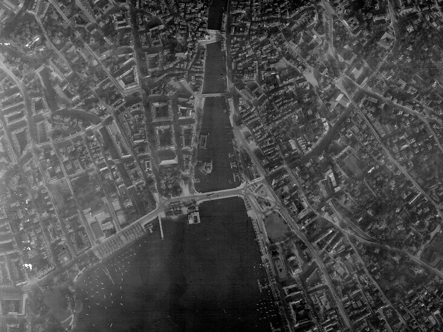A historic black and white vertical aerial photo of a large city straddling a river. 3 bridges cross the river, which runs into a lake at the bottom edge of the image. The city's streets and buildings are clearly visible.