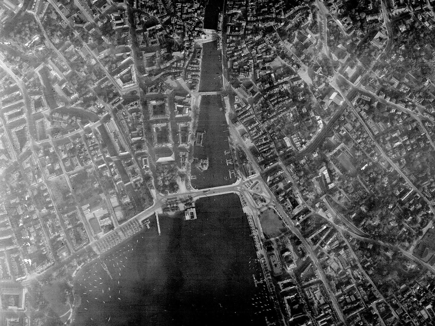 A historic black and white vertical aerial photo of a large city straddling a river. 3 bridges cross the river, which runs into a lake at the bottom edge of the image. The city's streets and buildings are clearly visible.