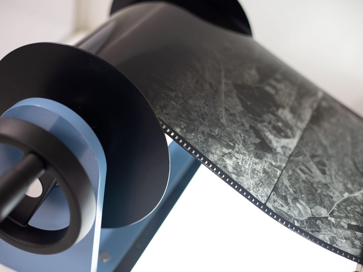 A blue support for large rolls of film with a black winding handle. A black and white aerial film is being supported on the winder. It is lit from below with a white light.