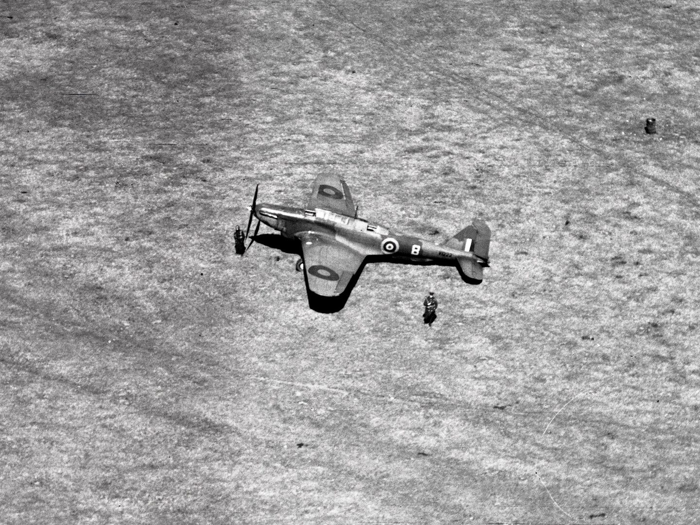 A historic black and white oblique aerial photo of a Fairey Battle aircraft with RAF roundels on each wing stationary in the middle of a large field. Two figures in uniform stand next to the aircraft.