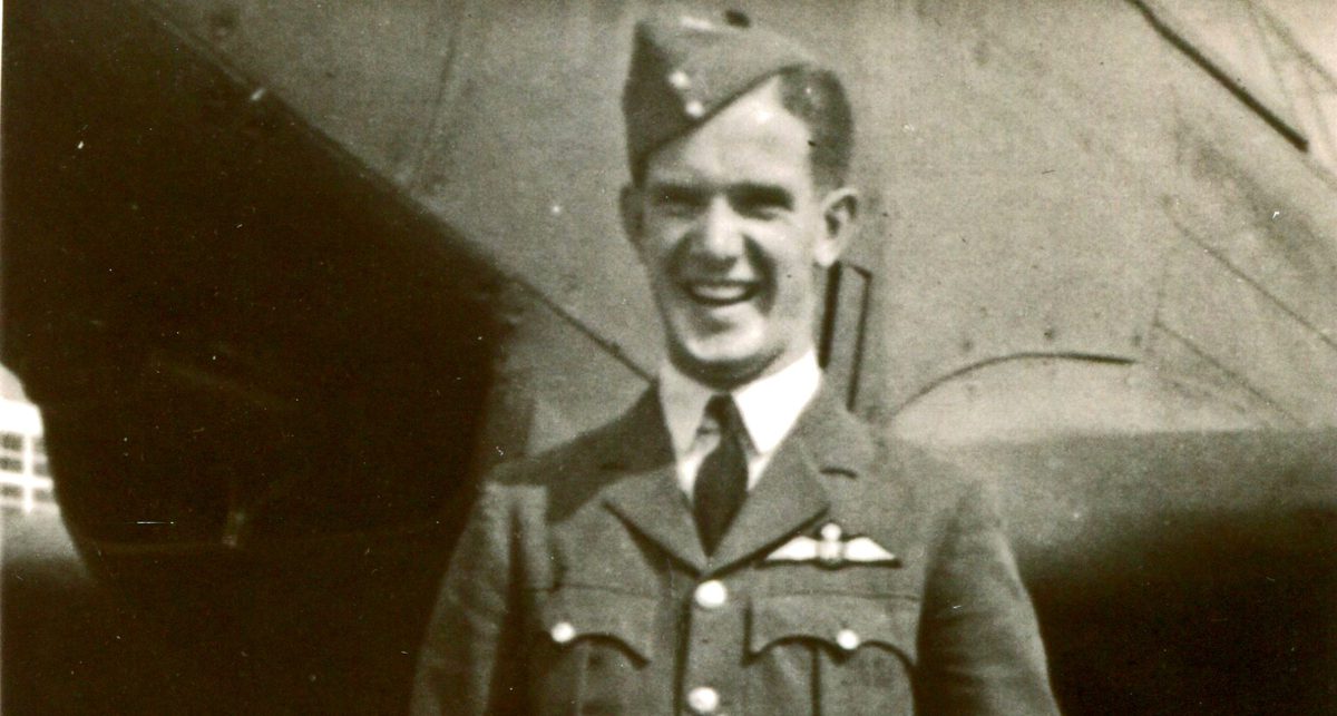 A historic sepia-coloured photo of a smiling man in uniform. He wears a beret and has an RAF wings badge on his left breast. He is standing in front of an aircraft.