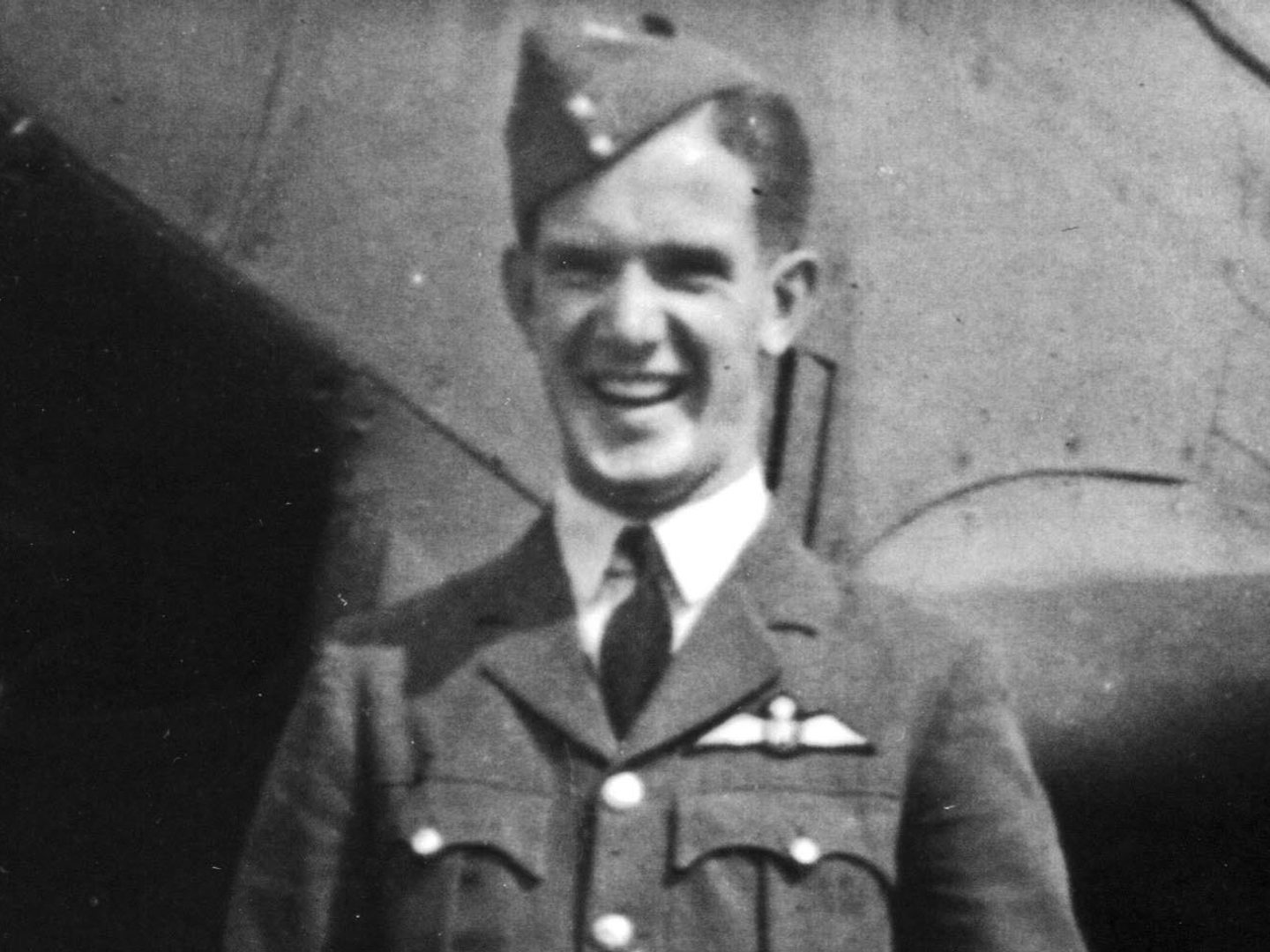 A historic sepia-coloured photo of a smiling man in uniform. He wears a beret and has an RAF wings badge on his left breast. He is standing in front of an aircraft.