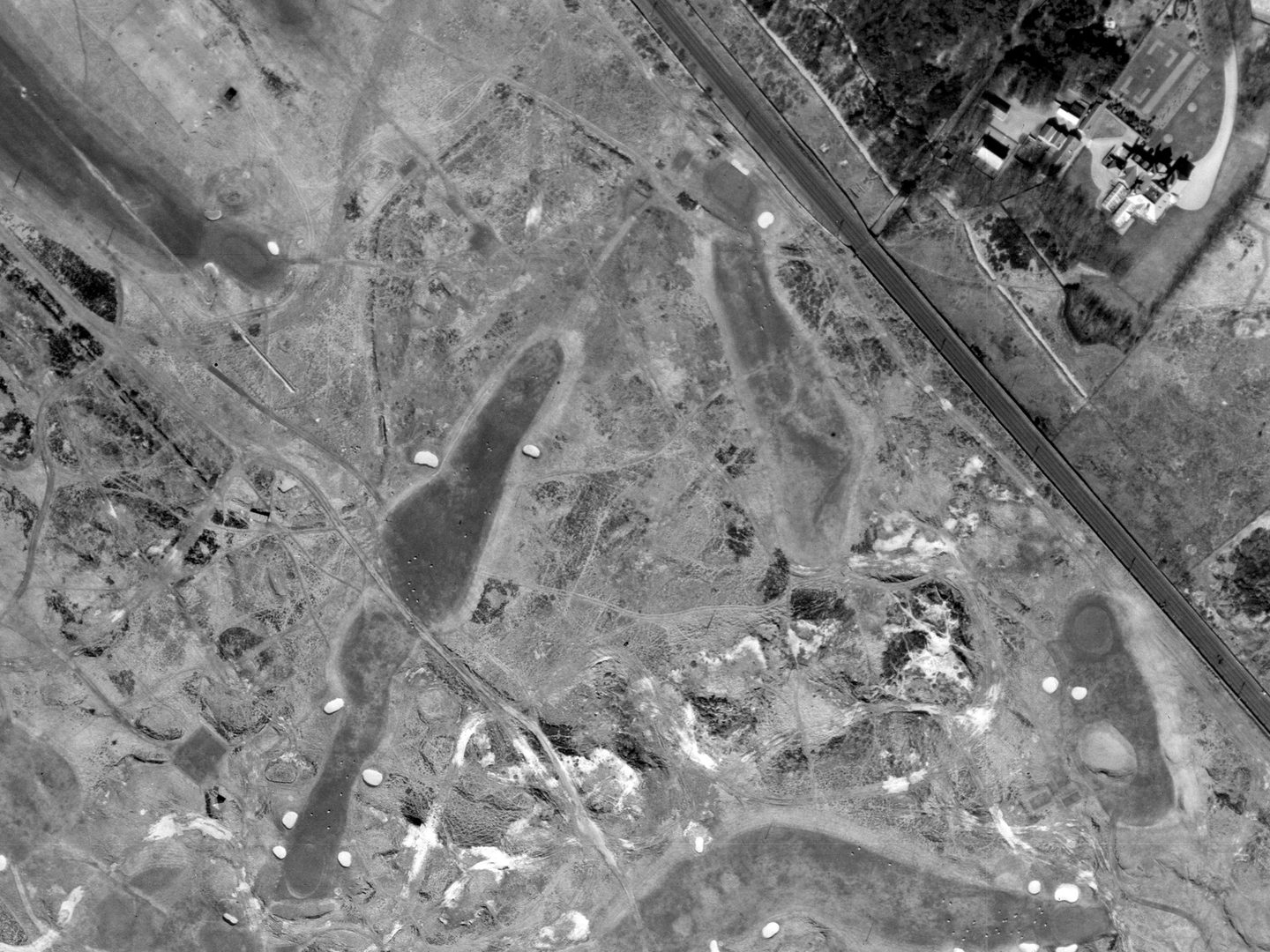 A historic black and white vertical aerial photo of a golf course with tees, greens and bunkers, with focus on a fairly short, straight hole running from top centre to bottom left.