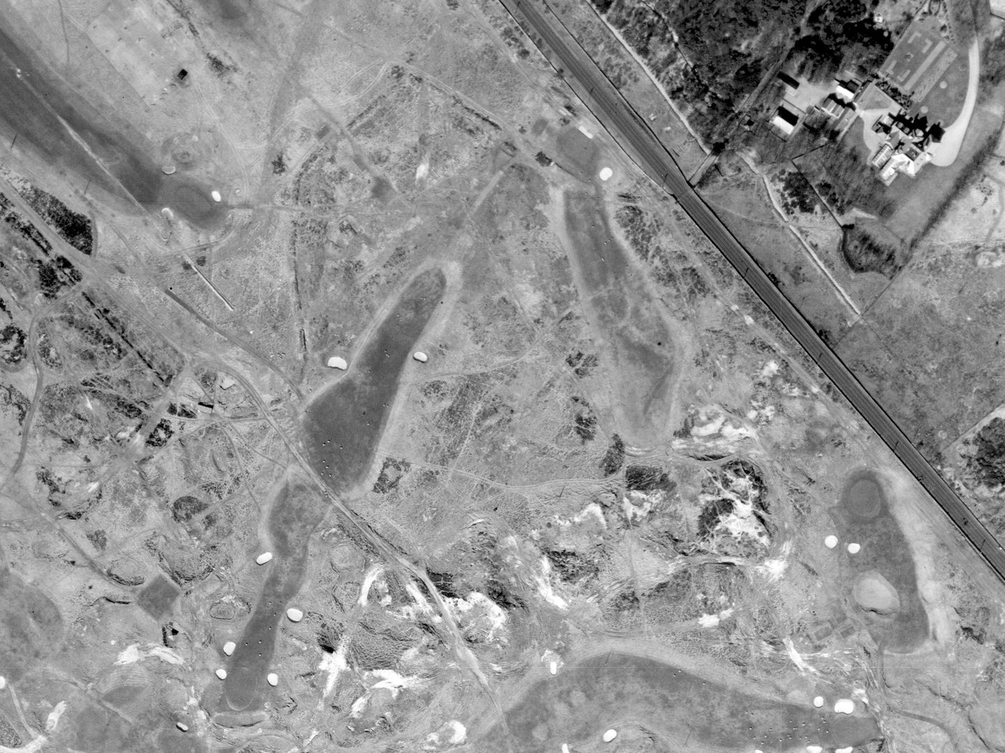 A historic black and white vertical aerial photo of a golf course with tees, greens and bunkers, with focus on a fairly short, straight hole running from top centre to bottom left.