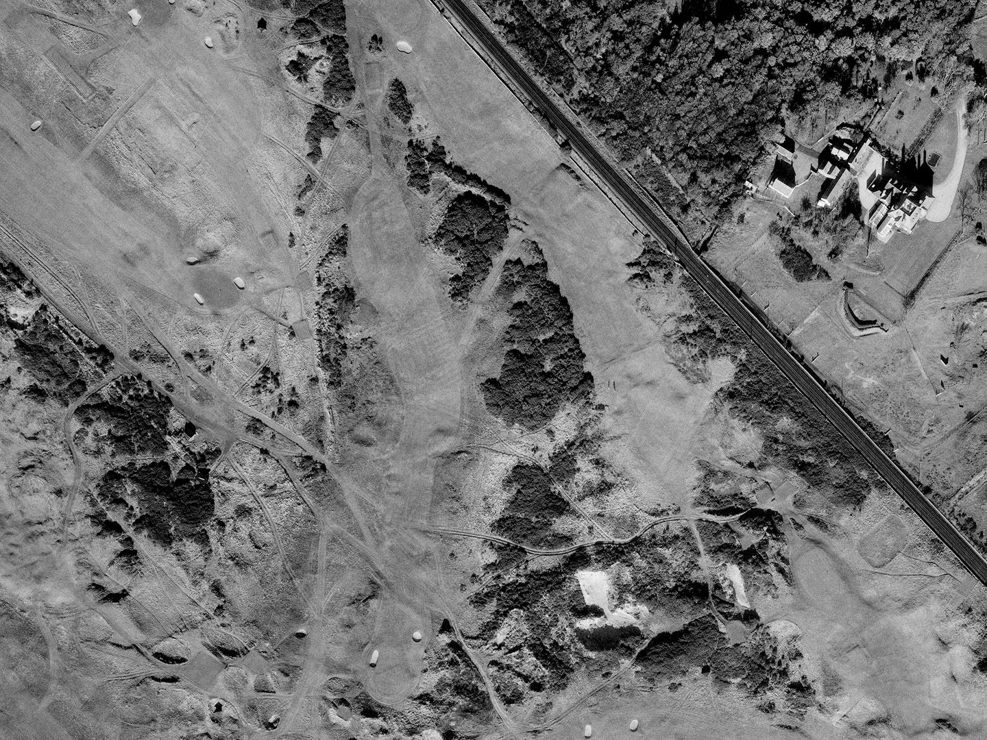 A historic black and white vertical aerial photo of a golf course with tees, greens, bunkers and gorse bushes, with focus on a medium-length, kinked hole running from top centre to bottom left.