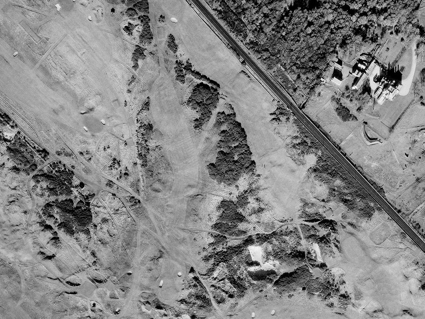A historic black and white vertical aerial photo of a golf course with tees, greens, bunkers and gorse bushes, with focus on a medium-length, kinked hole running from top centre to bottom left.