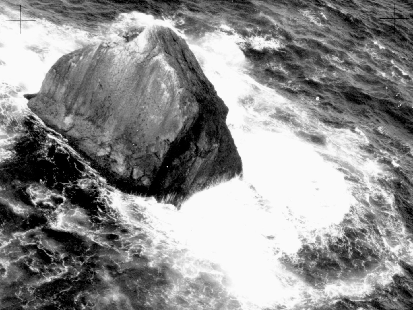 A historic black and white oblique aerial photo of Rockall - a triangular shaped rock surrounded by sea and the white foam of crashing waves.