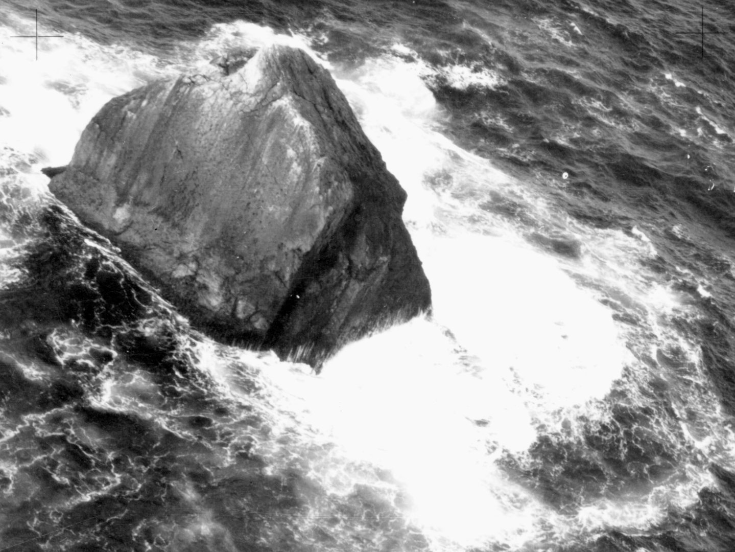 A historic black and white oblique aerial photo of Rockall - a triangular shaped rock surrounded by sea and the white foam of crashing waves.