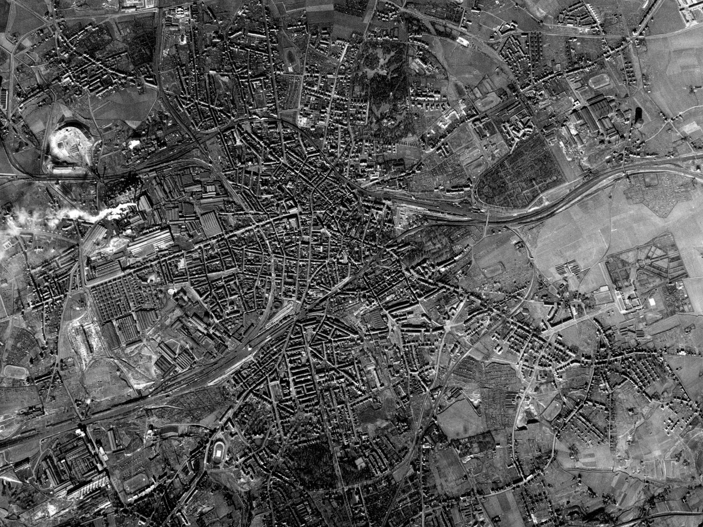 A historic black and white vertical aerial photo of a large town surrounded by farm fields, factories and industrial areas.