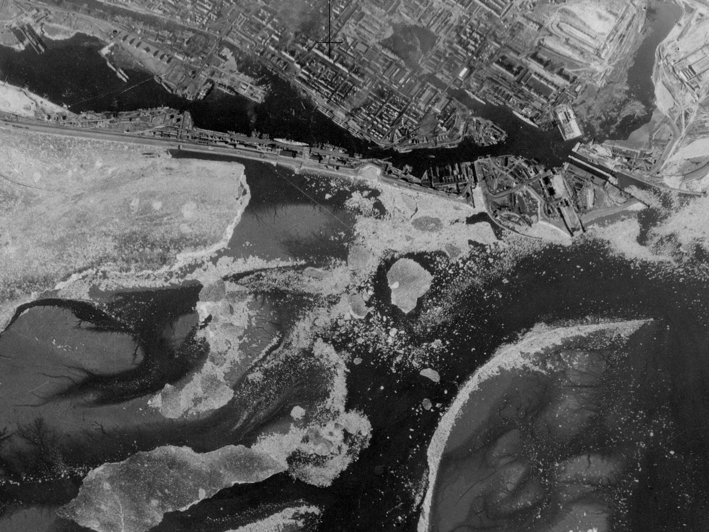 A historic black and white vertical aerial photo of a town on the shore of a sea. The town is along the top edge of the photo, with the sea below it, interspersed with what might be ice or sandbars. A spit of land sandwiches an area of water between it and the town. There are docks on the spit of land with ships berthed there.