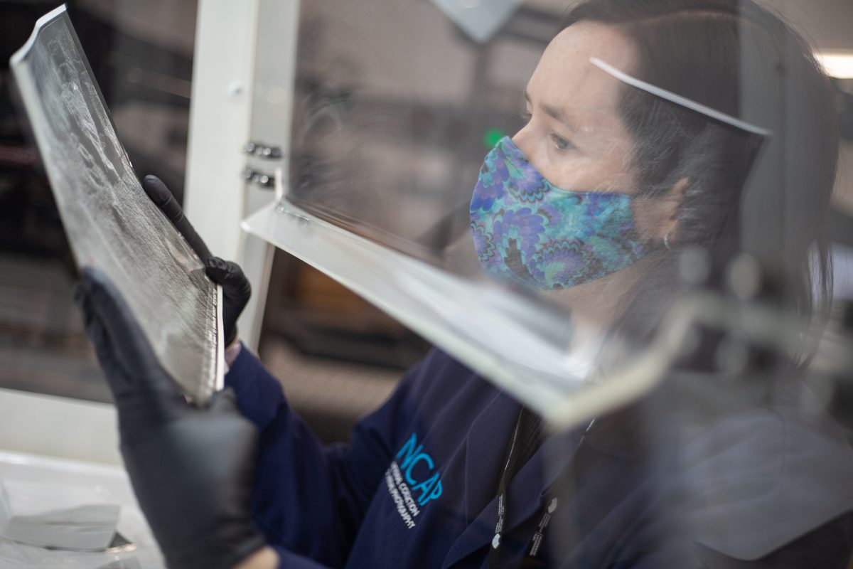 A member of NCAP staff in a blue lab coat and wearing a patterned face mask examines a black and white photographic print within a fume hood.
