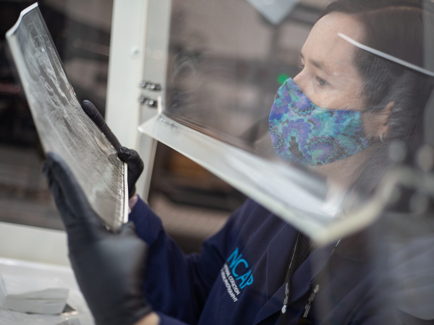 A member of NCAP staff in a blue lab coat and wearing a patterned face mask examines a black and white photographic print within a fume hood.