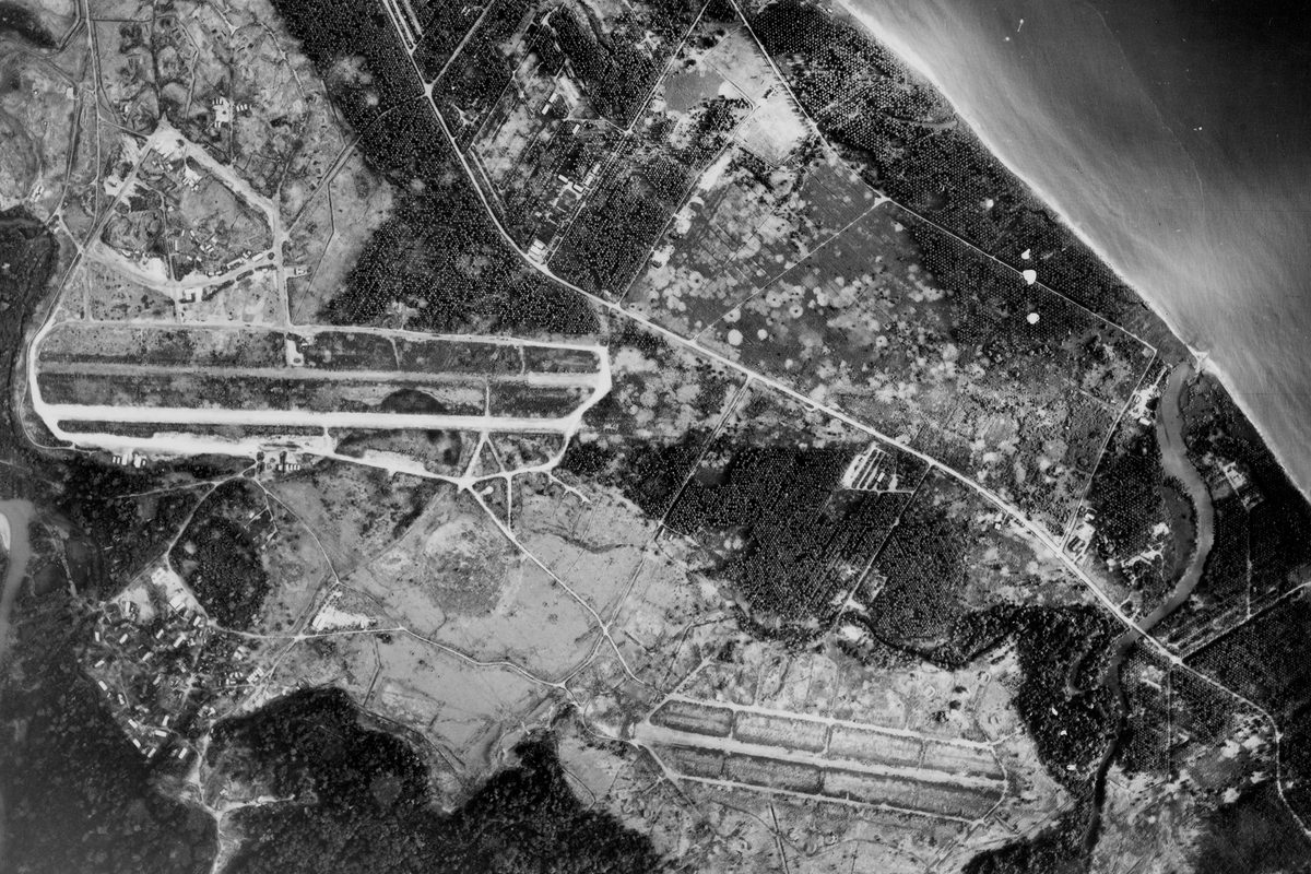 A historic black and white vertical aerial photo of an airport. 2 runways are visible running across the image separated by fields. A road runs diagonally from the top left to the bottom right of the image. Above the road is a settlement and coastline, with the sea visible in the top right corner.