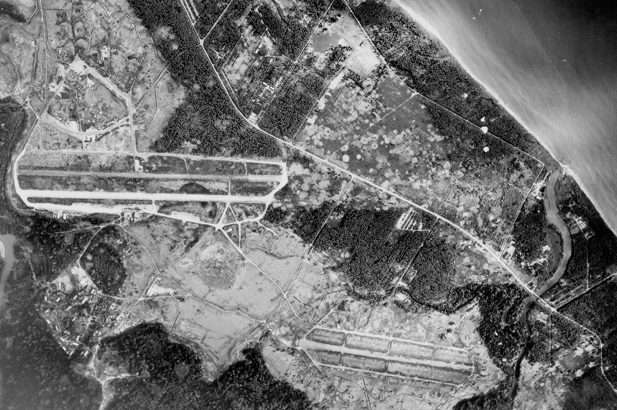 A historic black and white vertical aerial photo of an airport. 2 runways are visible running across the image separated by fields. A road runs diagonally from the top left to the bottom right of the image. Above the road is a settlement and coastline, with the sea visible in the top right corner.