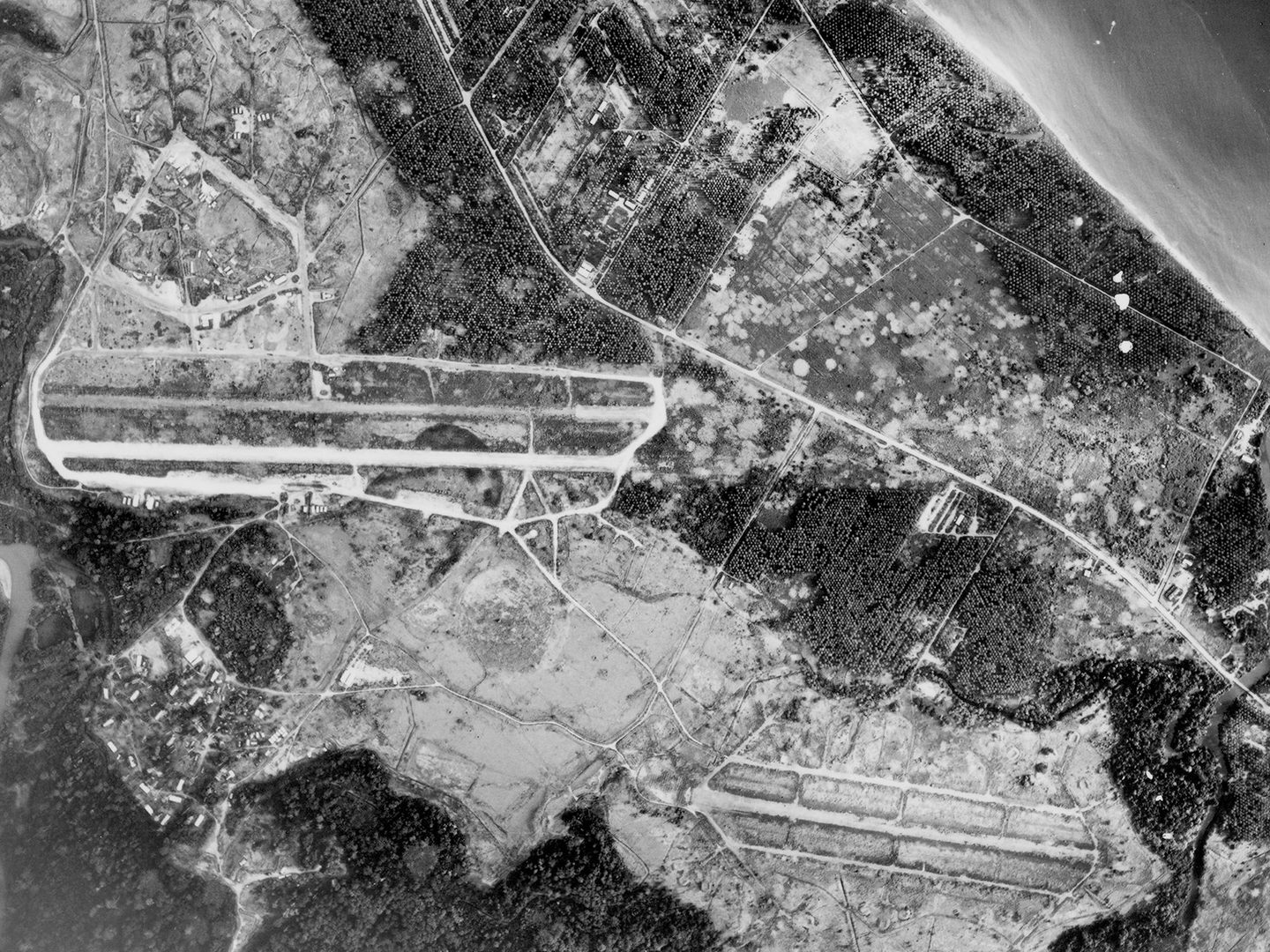 A historic black and white vertical aerial photo of an airport. 2 runways are visible running across the image separated by fields. A road runs diagonally from the top left to the bottom right of the image. Above the road is a settlement and coastline, with the sea visible in the top right corner.