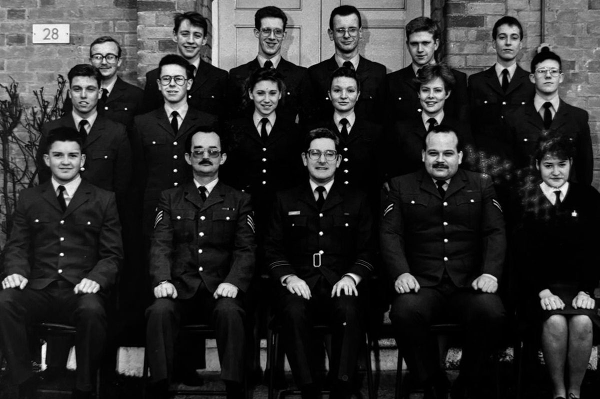 A historic black and white photo of 17 men and women un uniform, arranged in 3 rows in front of a brick building, with the first row seated.