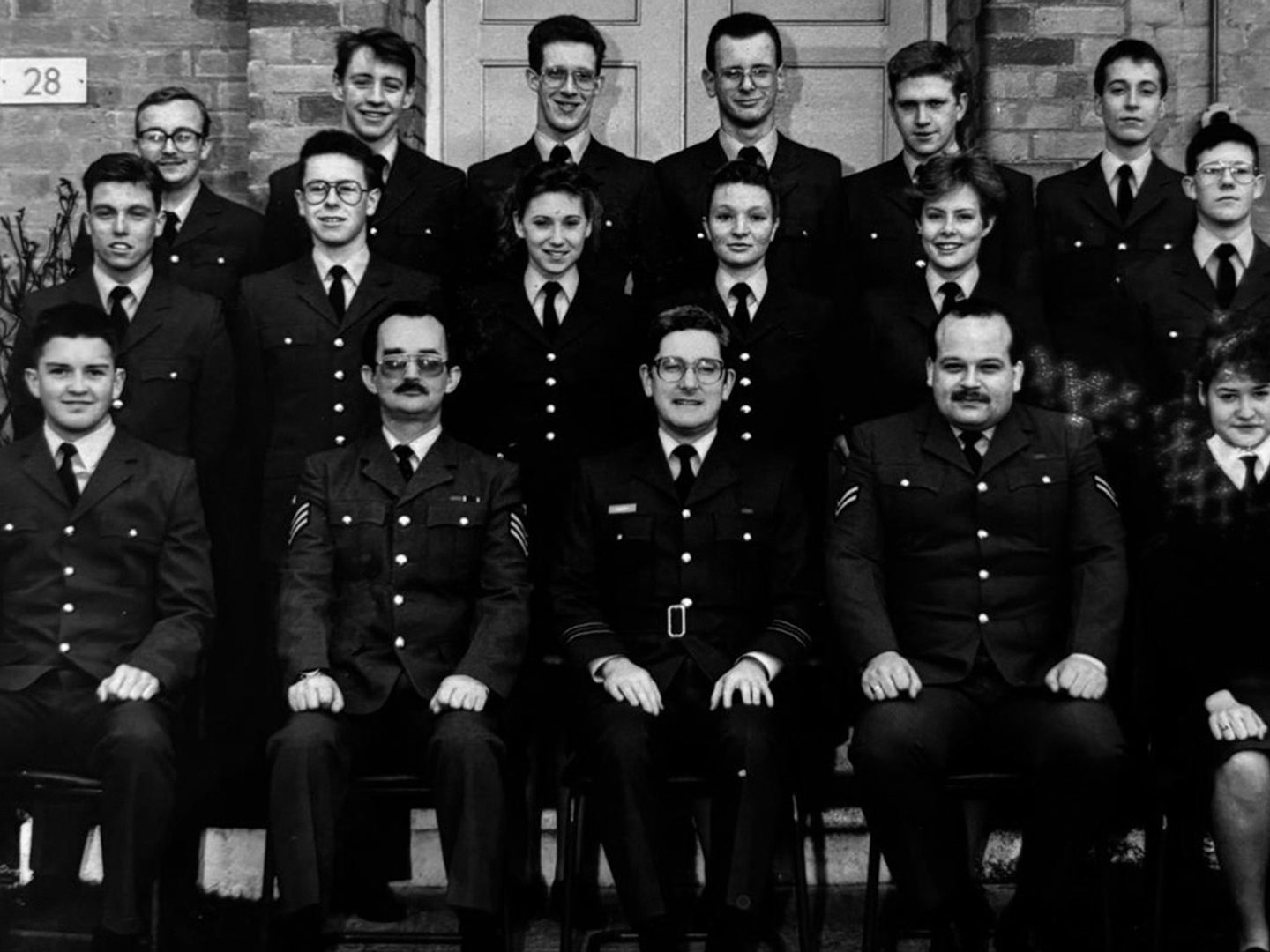 A historic black and white photo of 17 men and women un uniform, arranged in 3 rows in front of a brick building, with the first row seated.