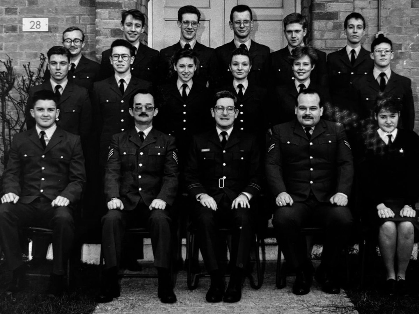 A historic black and white photo of 17 men and women un uniform, arranged in 3 rows in front of a brick building, with the first row seated.