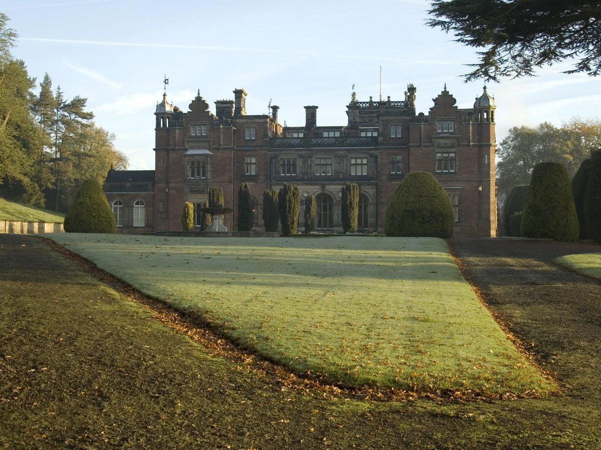 A grand red brick building with cupulas at each corner. In front of it is a lawn scattered with fallen leaves and lit by raking sunshine. Trees are visible to either side of the building, and there is a row of yew trees between it and the lawn.