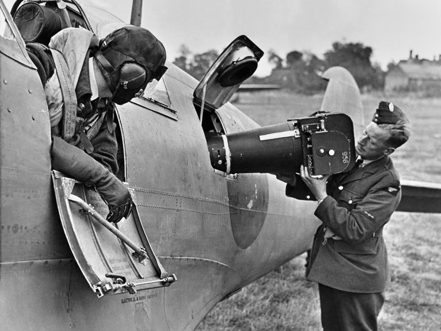 A black and white historic photo of a man in uniform loading a large camera into the side of a plane. A second man in a flying helmet is leaning out of the plane watching him.