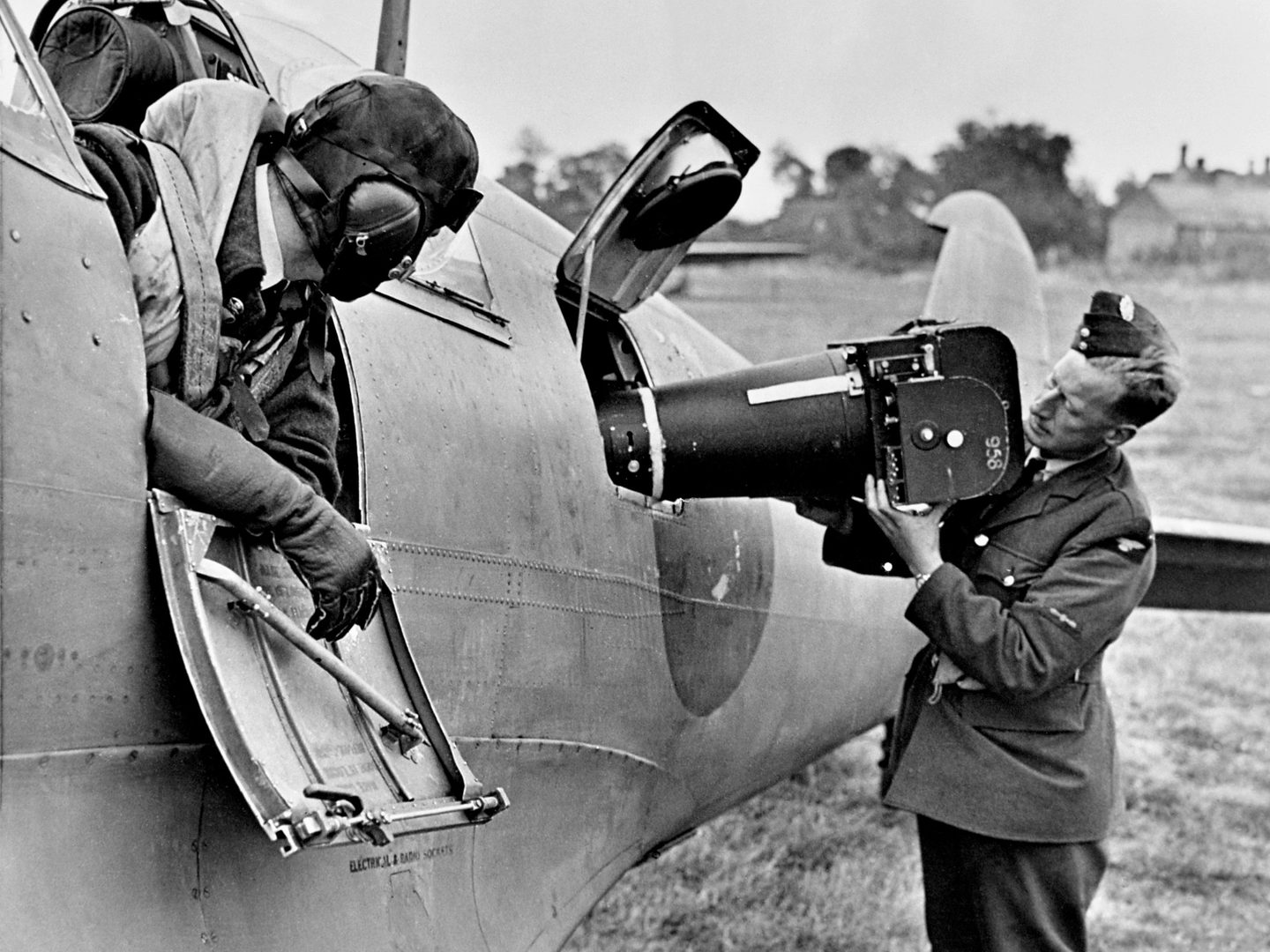 A black and white historic photo of a man in uniform loading a large camera into the side of a plane. A second man in a flying helmet is leaning out of the plane watching him.