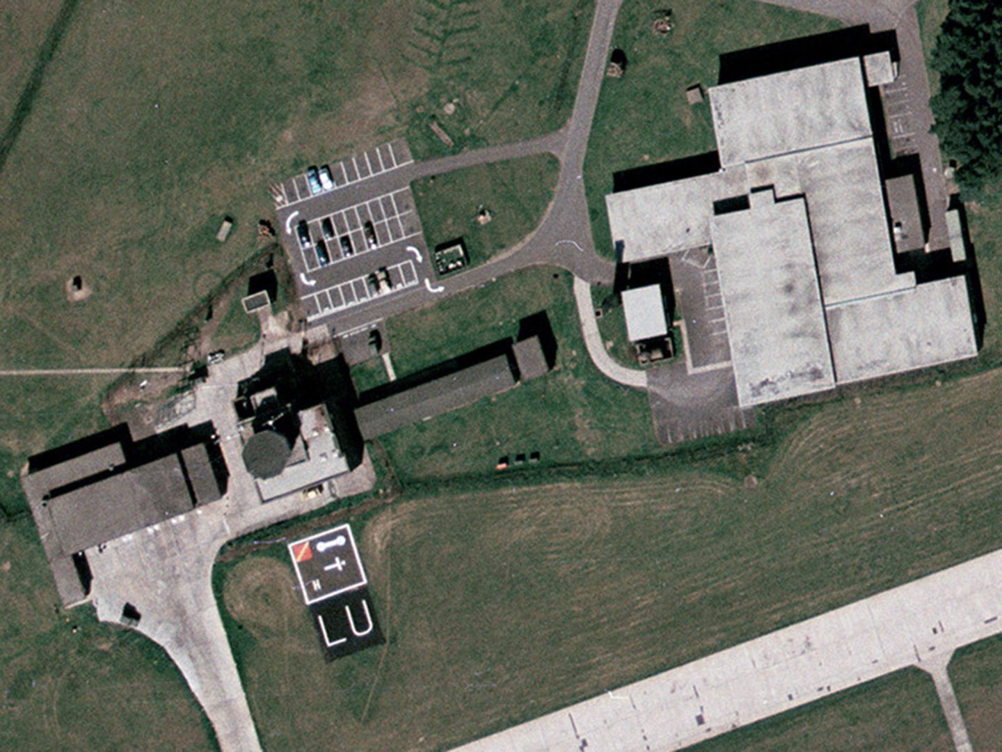 A historic colour vertical aerial photo of an area of an airfield. To the right is a large building with a light grey flat roof surrounded by grass. To the left are green-roofed buildings, a car park and an octagonal tower. In front of the tower are several symbols arranged within a square and the letters 'LU'.