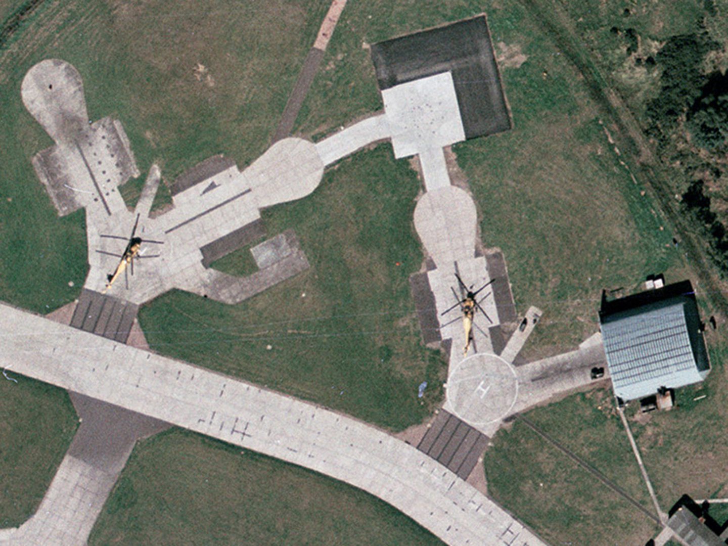 A historic colour vertical aerial photo of 2 yellow helicopters parked on light-coloured concrete. A light-coloured concrete road runs along the bottom of the image, and a grey-roofed building is to the right.
