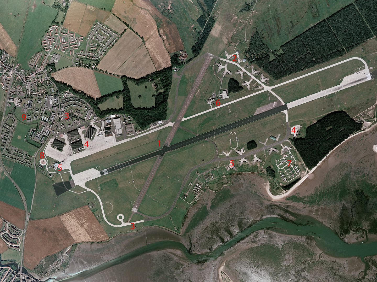 A historic colour vertical aerial photo of an airfield. The main runway runs across the image from right to left, with a shorter runway crossing it at a diagonal towards the left-hand end. A white road runs parallel to and above the main runway. At the left-hand end of the road are 4 large hangars. A village is located in the top left-hand corner of the image, just above the hangars. Red numbers indicate points of interest.