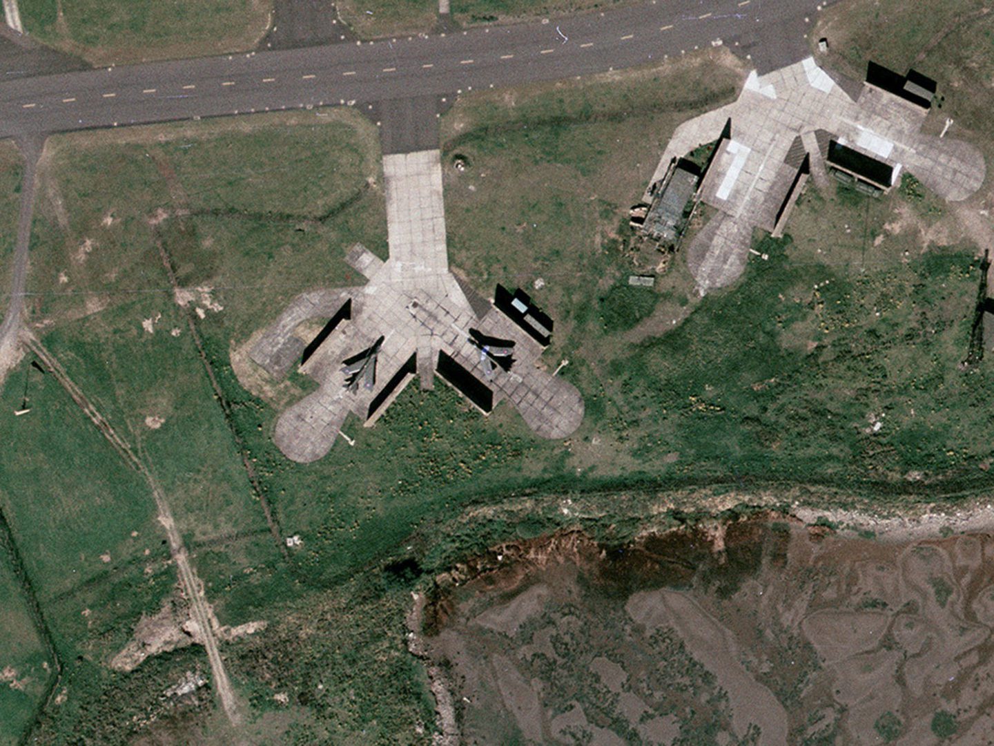 A historic colour vertical aerial photo of two aircraft parked on an area of light-coloured conrete, surrounded by grass. Each aircraft is flanked by a thick wall. A grey road runs along the top edge of the image.