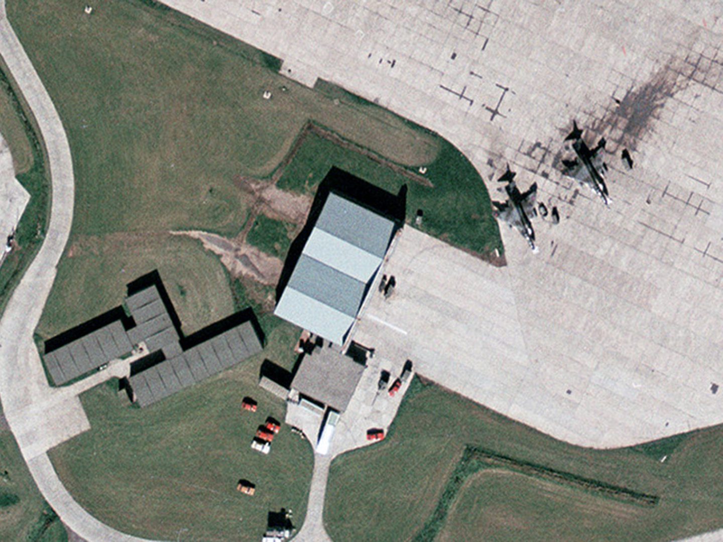 A historic colour vertical aerial photo of an area of an airfield. Prominently in the centre is a building with a light grey, pitched roof. Below it and to the left is grass, to the right is light-coloured concrete. 2 aircraft painted in camouflage are parked on the concrete. Further buildings with green roofs are to the left.