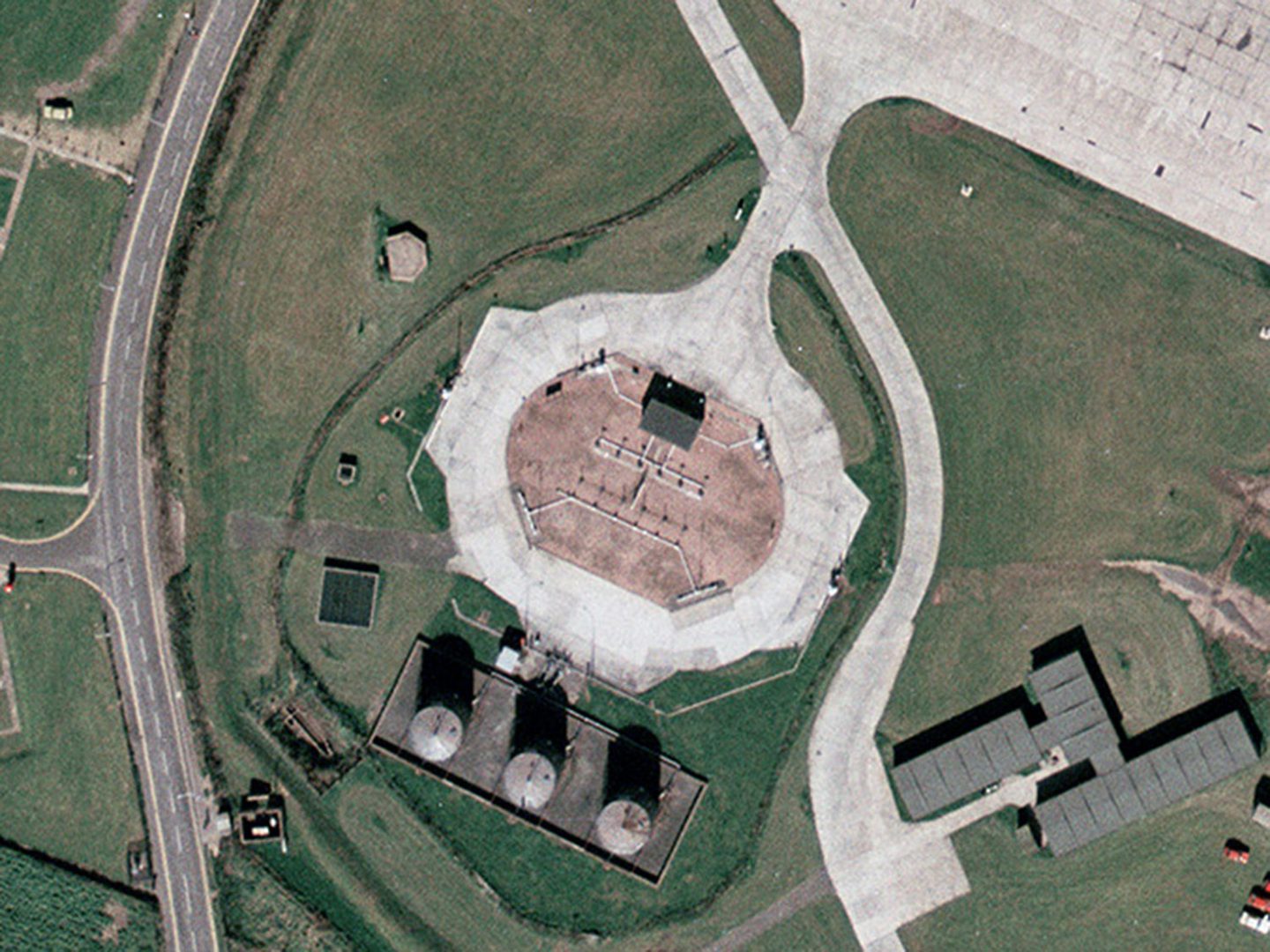 A historic colour vertical aerial photo of a fuel storage area. In the centre of the image is the top of the underground storage with pipe inlets just visible. At the bottom centre of the image are 3 large fuel storage tanks. The storage area is surrounded by grass, with a grey tarmac road to the left and other light-coloured concrete roads visible.