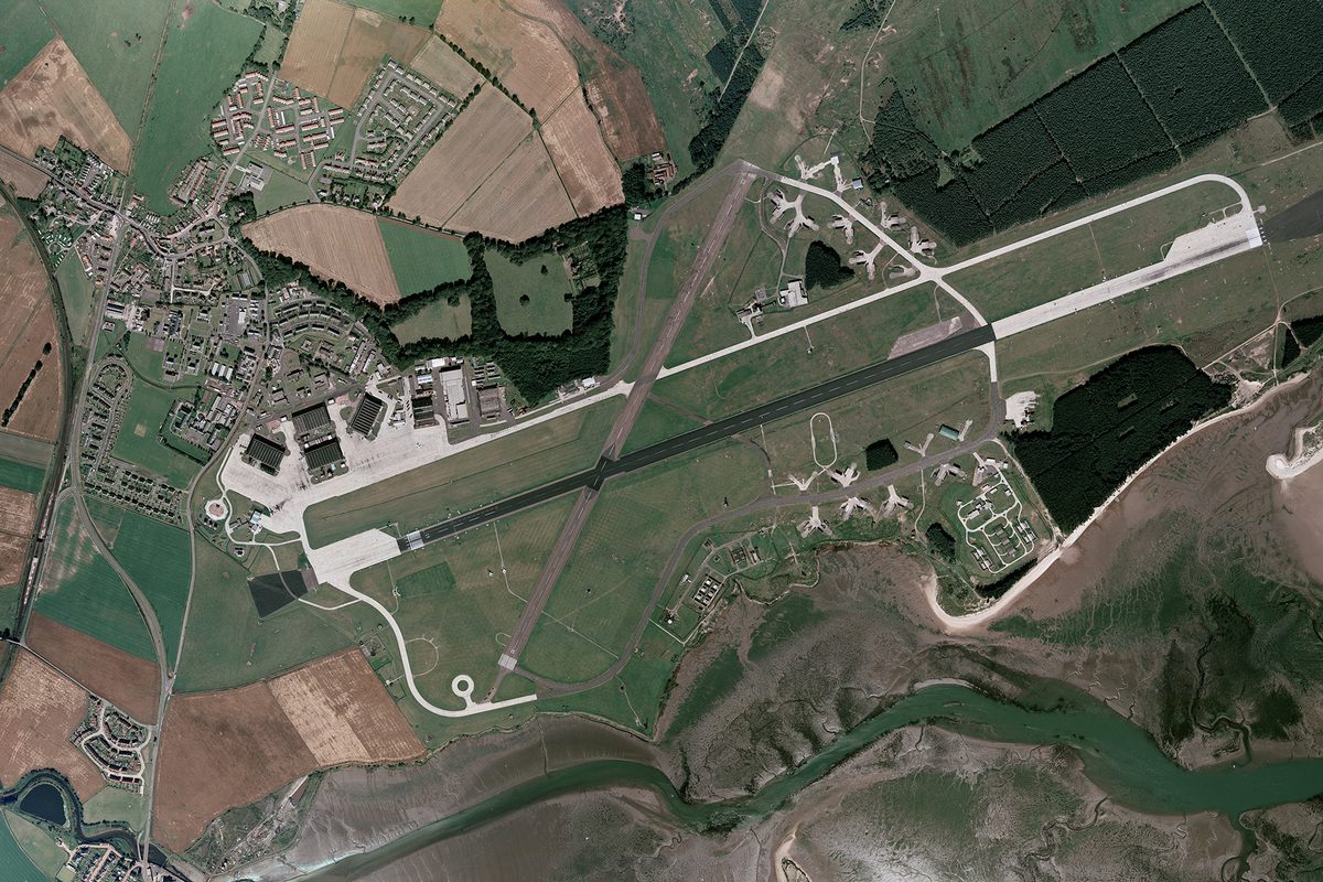 A historic colour vertical aerial photo of an airfield. The main runway runs across the image from right to left, with a shorter runway crossing it at a diagonal towards the left-hand end. A white road runs parallel to and above the main runway. At the left-hand end of the road are 4 large hangars. A village is located in the top left-hand corner of the image, just above the hangars.