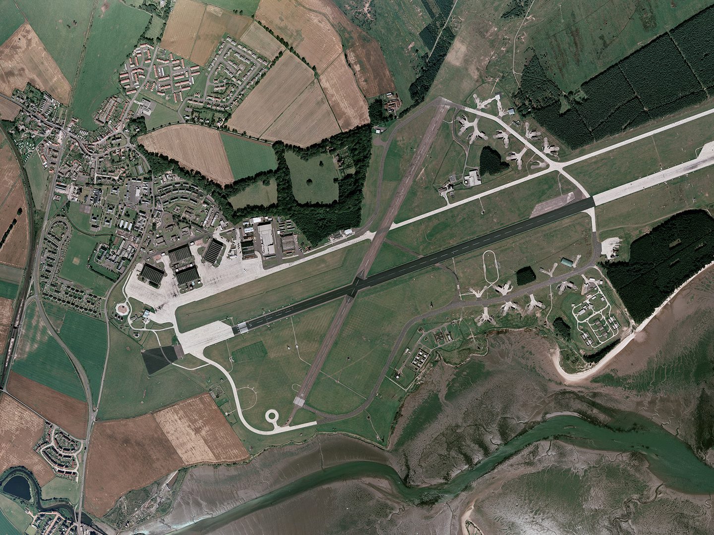 A historic colour vertical aerial photo of an airfield. The main runway runs across the image from right to left, with a shorter runway crossing it at a diagonal towards the left-hand end. A white road runs parallel to and above the main runway. At the left-hand end of the road are 4 large hangars. A village is located in the top left-hand corner of the image, just above the hangars.