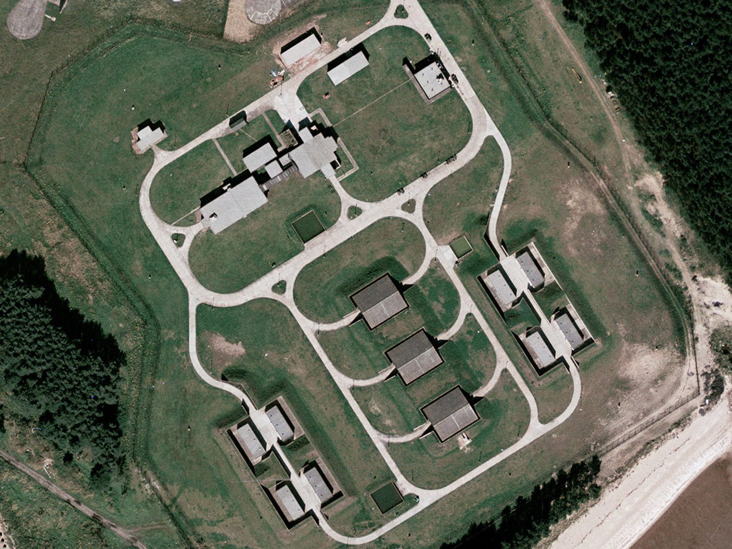 A historic colour vertical aerial photo of 9 low-roofed buildings arranged in a symmetrical pattern and linked by a regular pattern of roads. The 2 lines of a double fence are visible around the buildings, with areas of woodland outside the fence to the left and right.