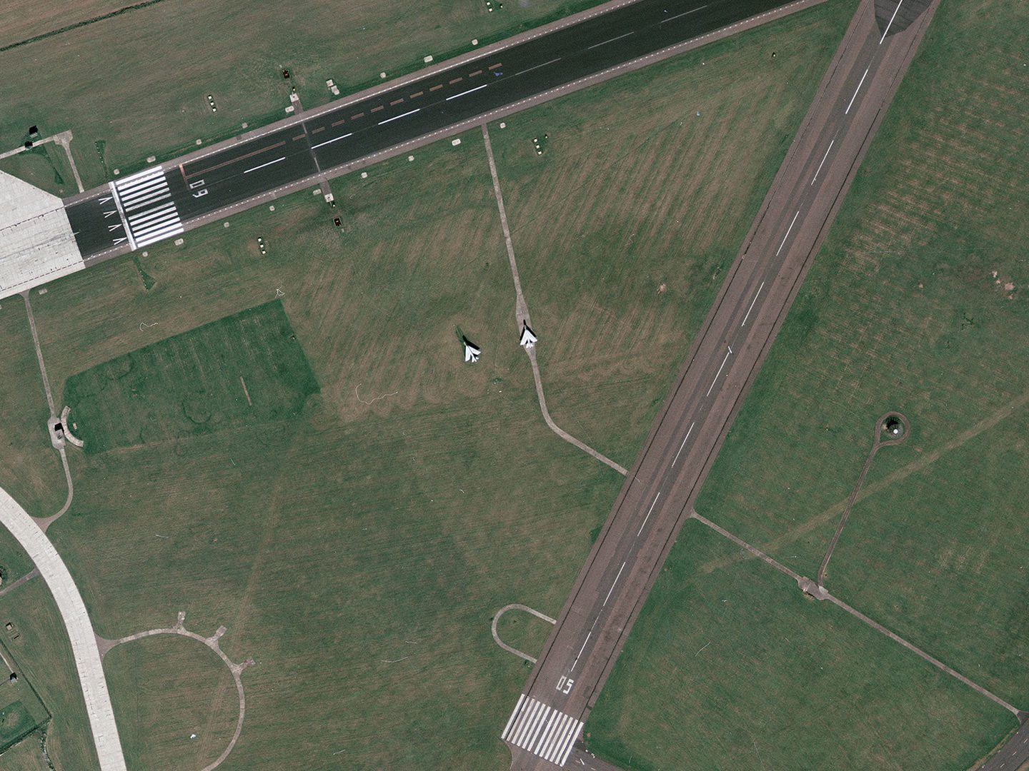 A historic colour vertical aerial photo of 2 runways running through grass. A dark grey runway runs along the top edge of the image, with a lighter grey one running from the centre bottom of the image towards the top right corner.